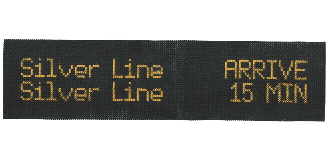 Cross-stitch of gold thread on black background, reading that a Silver Line bus is arriving, and another is coming in 15 minutes in appearance of the hanging LED schedules on arrival platforms